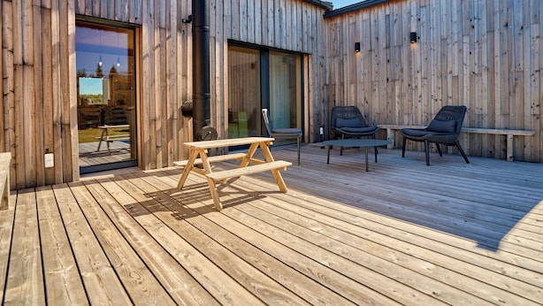 Eine Holzterrasse mit Picknicktisch und zwei Stühlen, einer schwarzen Rohrleitung und einer Glastür, die in einen Außenbereich führt.