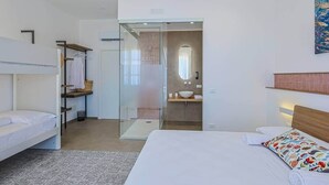 Ein modernes Hotelzimmer mit Stockbett, Duschkabine aus Glas, einem separaten Badezimmer inklusive Waschbecken und Spiegel sowie einem großen Bett mit farbigem Kissen.