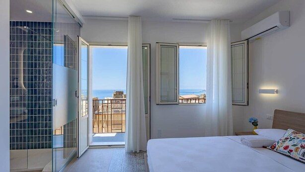 A hotel room with a bed, a glass-enclosed shower, a balcony with a view of the sea, and a large window. - Room Castel Perollo - Camera Verde by Biddizzi Rari - Historic Center in Sciacca (Sciacca)