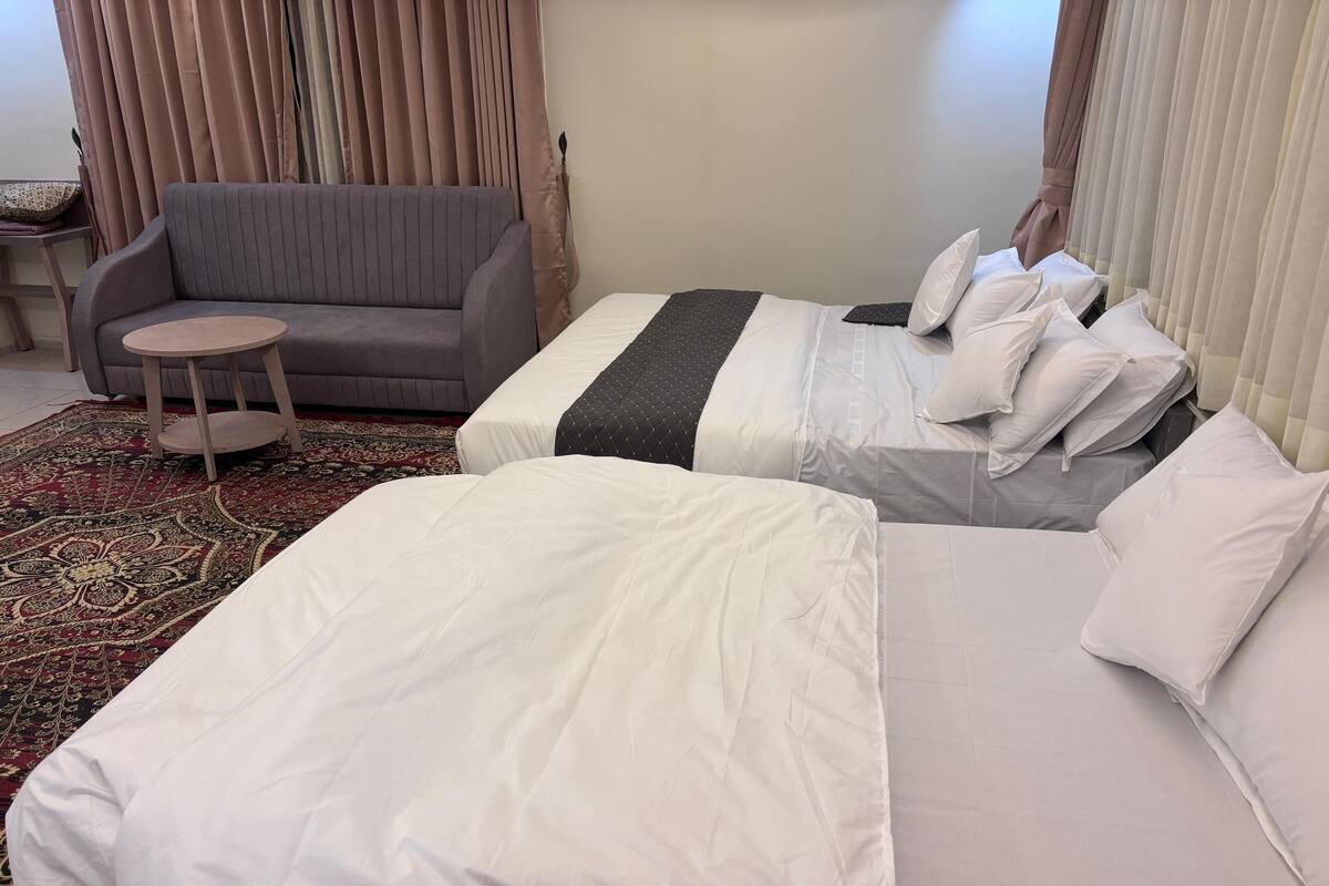 Habitació d'hotel amb dos llits, un sofà gris, una taula petita rodona i una catifa amb dibuixos.