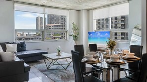 Een moderne woonkamer met een bank, eettafel en stoelen, grote ramen met uitzicht over de stad, en een plafond met zichtbare balken.