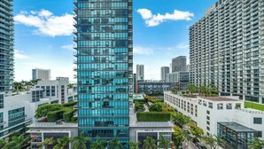 A modern high-rise building with a glass facade, surrounded by other high-rise buildings and palm trees. - Midtown Muse -Walkable Cozy amenity bldg w parking (Miami)