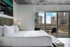 A hotel room with a large bed, a nightstand with a lamp, a view of high-rise buildings through large windows. - Midtown Muse -Walkable Cozy amenity bldg w parking (Miami)