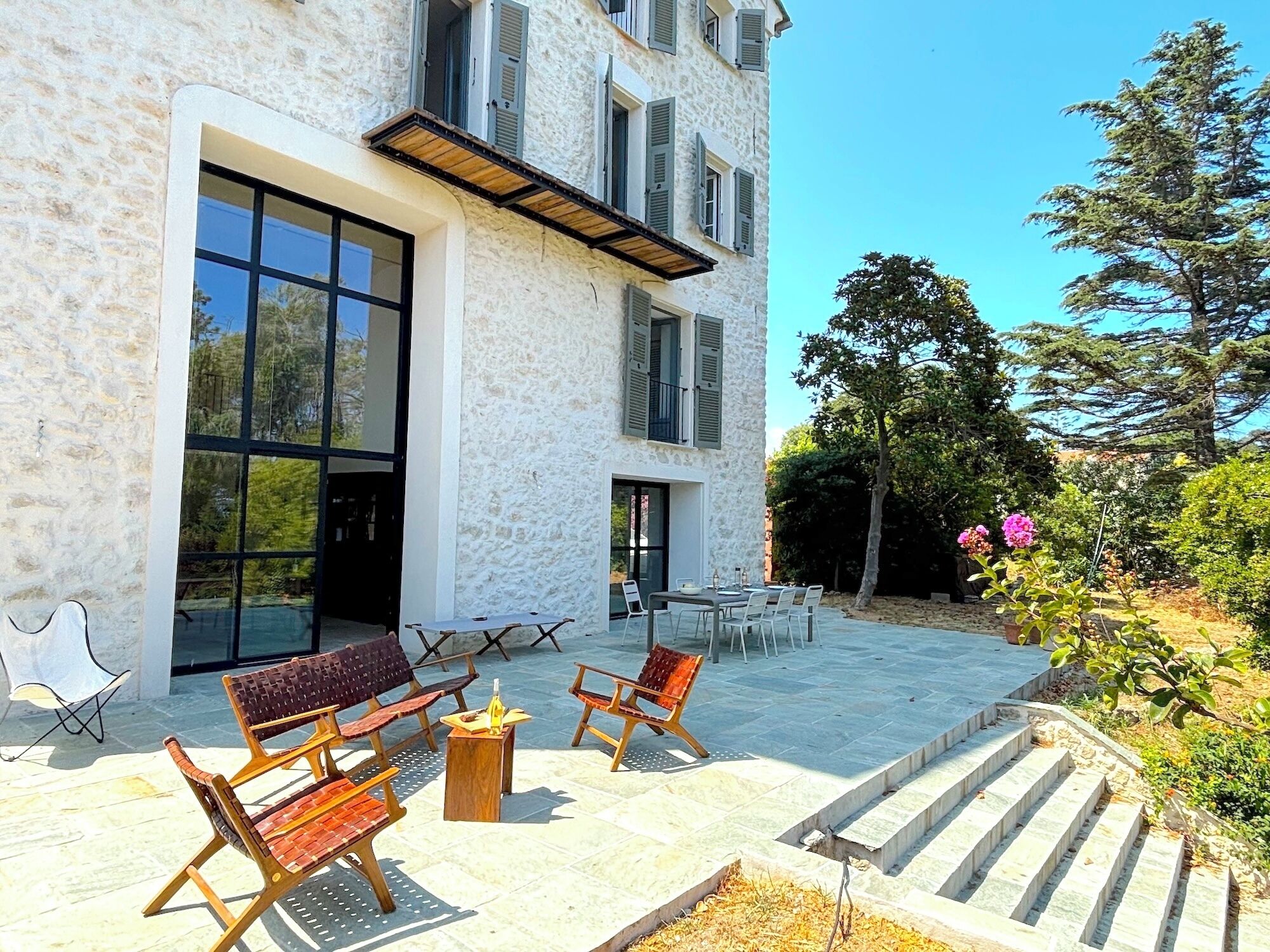 A stone building with a glass door and windows, outdoor seating with benches and a table, and a staircase leading to a garden area.