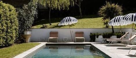 A swimming pool with lounge chairs and umbrellas, surrounded by greenery and a building in the background.