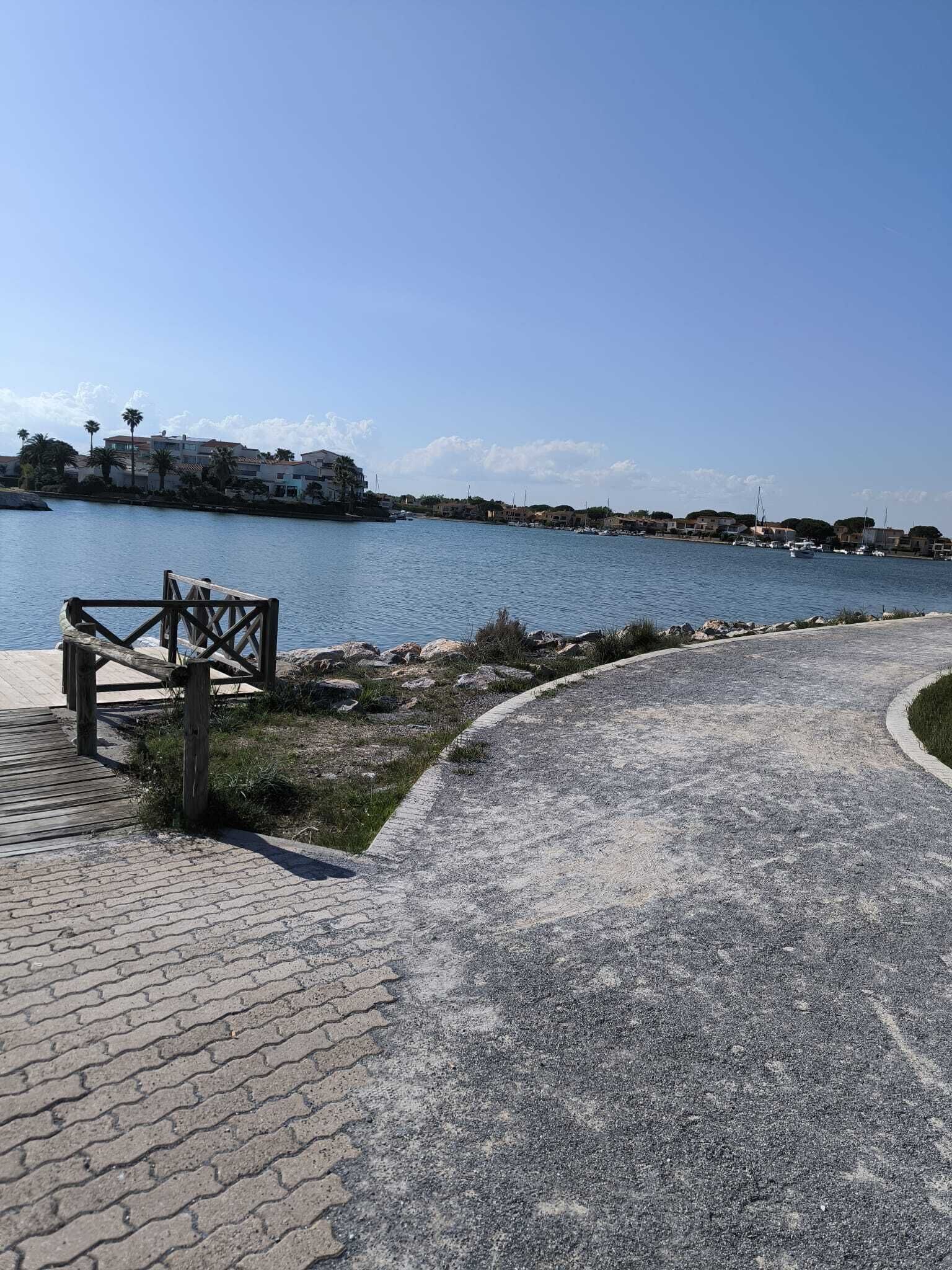 Eine Uferpromenade mit Holzlattenzaun, gepflastertem Weg und Blick auf Gebäude gegenüber dem Wasser.