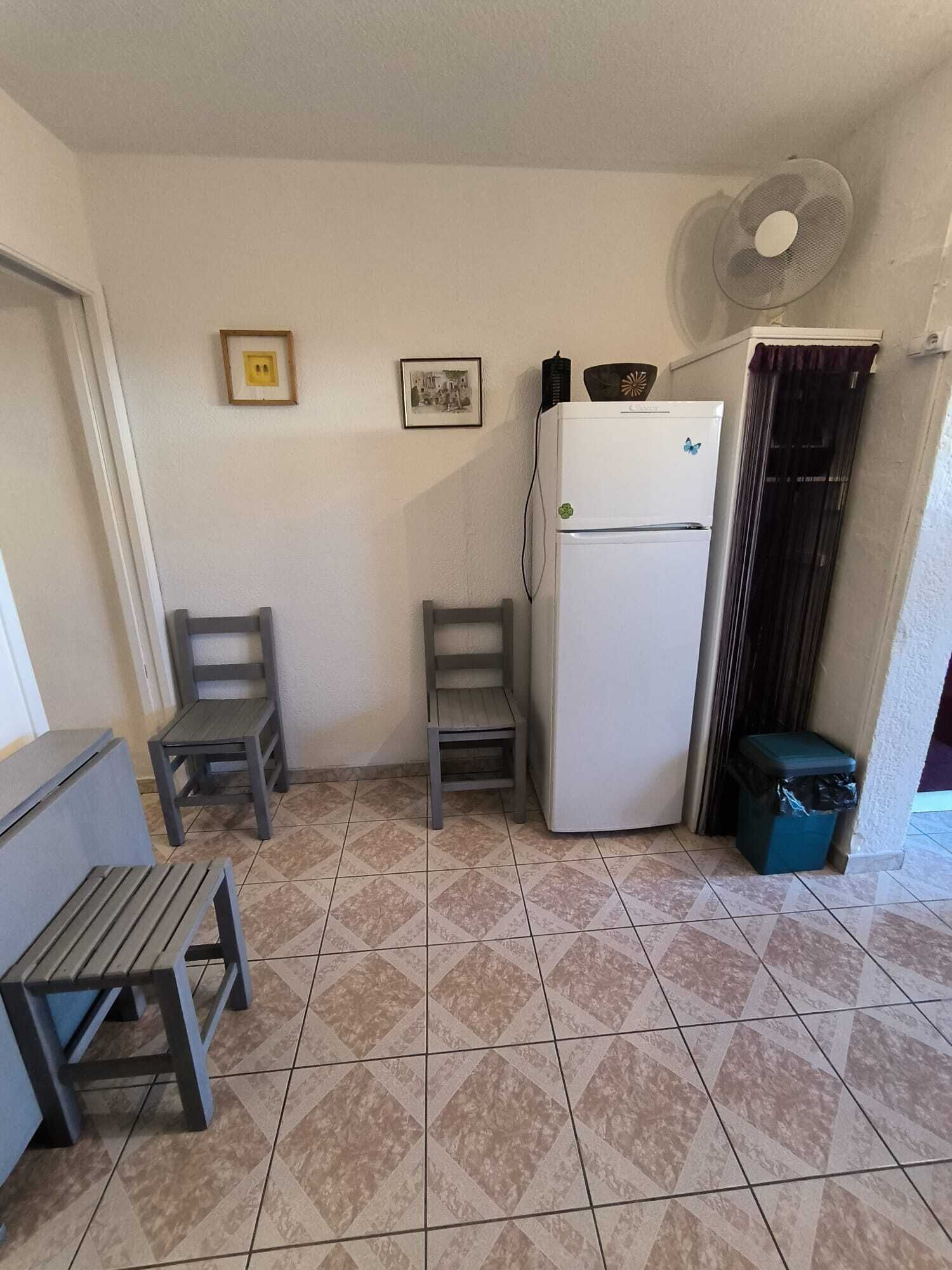 Een kleine keuken met een koelkast, twee stoelen, een bankje, een prullenbak en een ventilator aan de muur.
