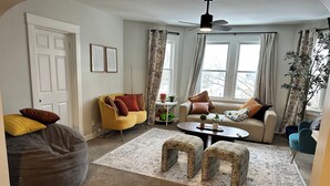 Ett vardagsrum med en gul fåtölj, en beige soffa och ett soffbord. Det finns två små pallar, en sittpuff och en takfläkt.