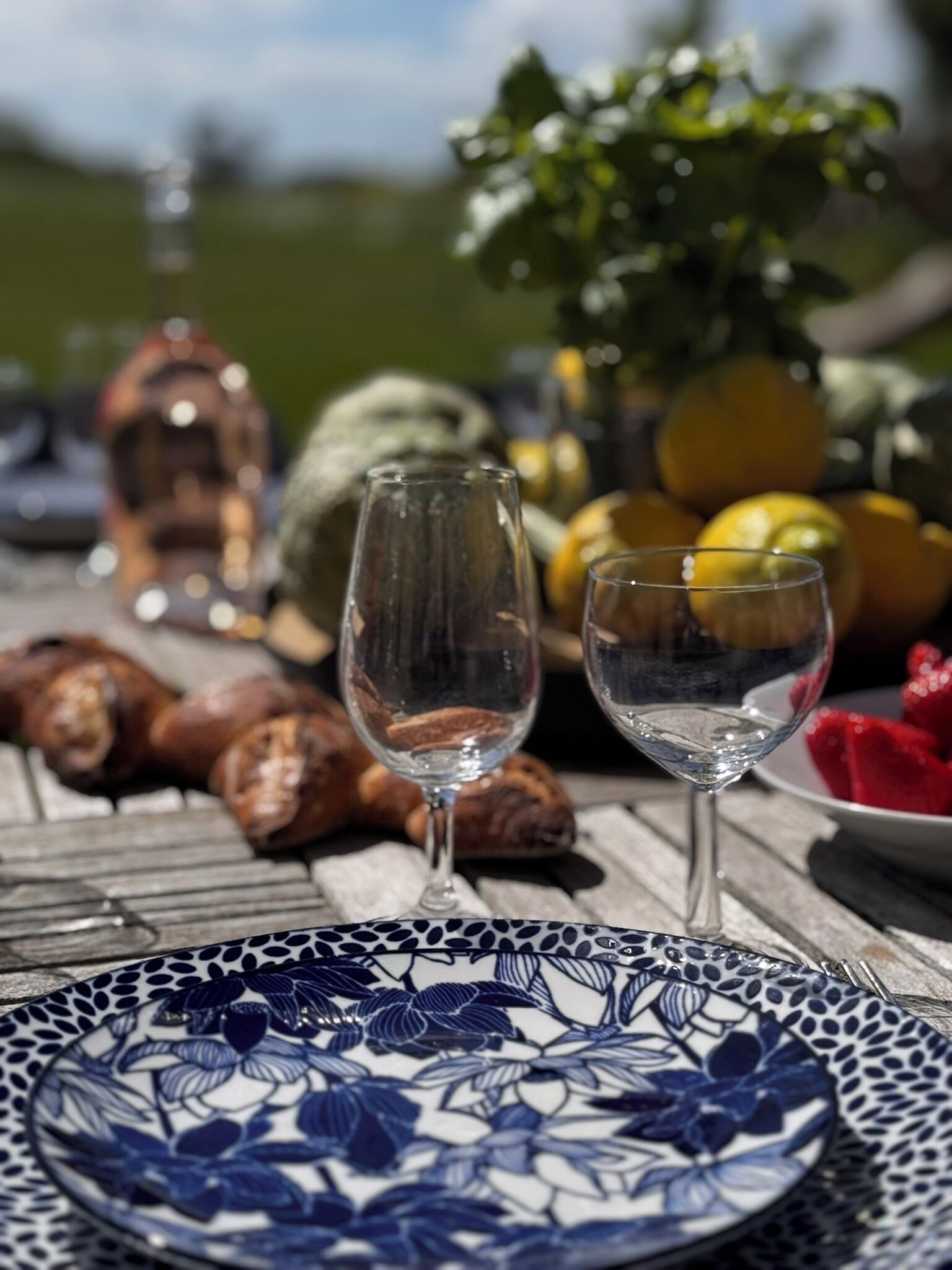 Une table avec deux verres à vin, une assiette ornée de motifs floraux et divers aliments, notamment du pain et des fruits.