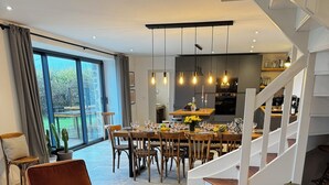 Een moderne woonkamer met een bruine leren bank, een houten salontafel met drankjes en bloemen, een eethoek met een grote tafel en stoelen, en een trap die naar een hoger gelegen verdieping leidt.