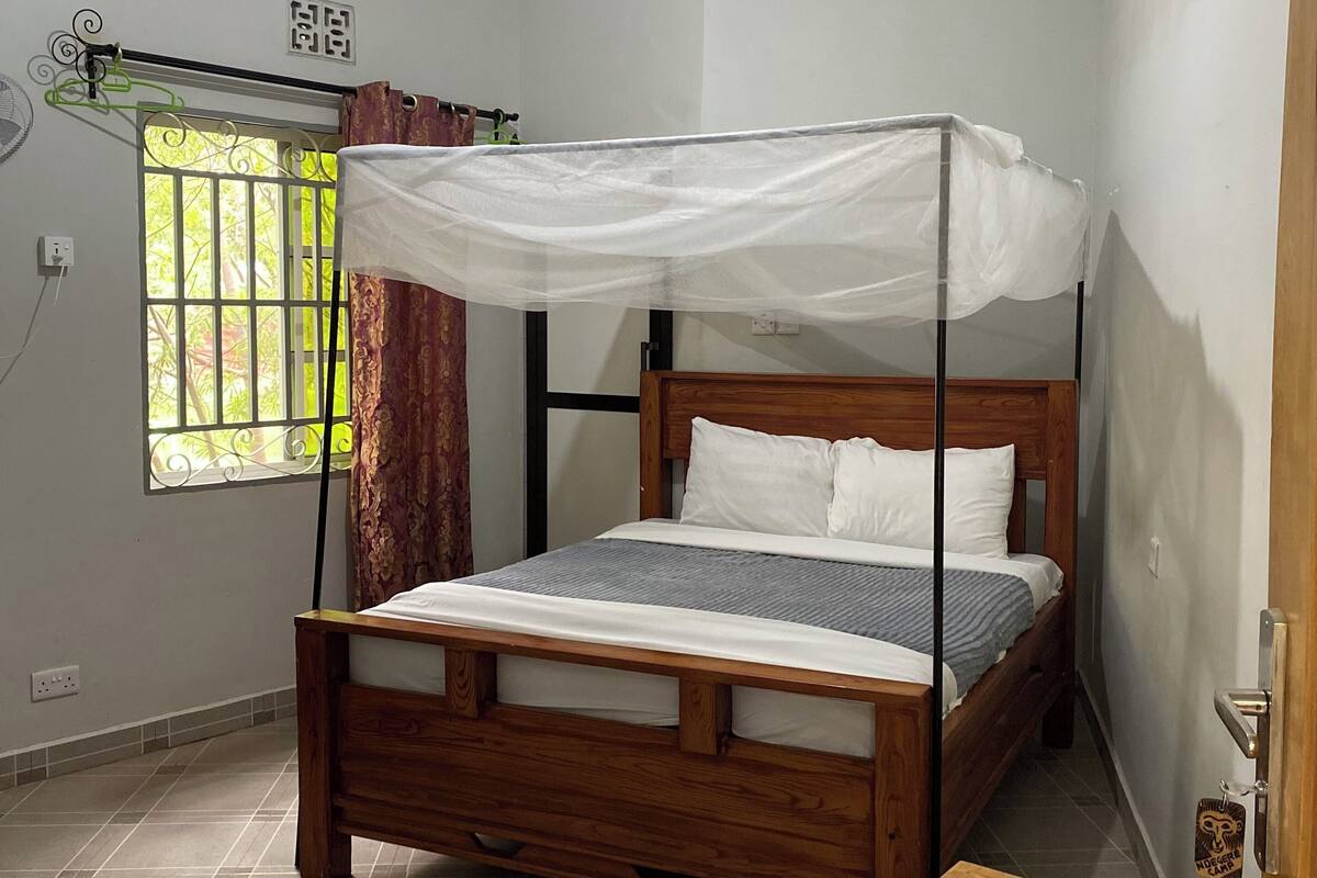 Un dormitorio con una cama de madera, mosquitero, una pequeña mesa de madera y una ventana con barrotes y cortinas.