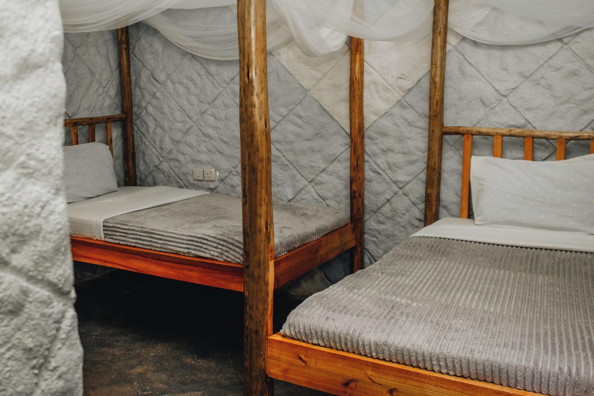 Una habitación con dos camas de madera, cada una con un colchón blanco y una manta gris. Las paredes están cubiertas con una tela acolchada, y hay un dosel blanco sobre una de las camas.