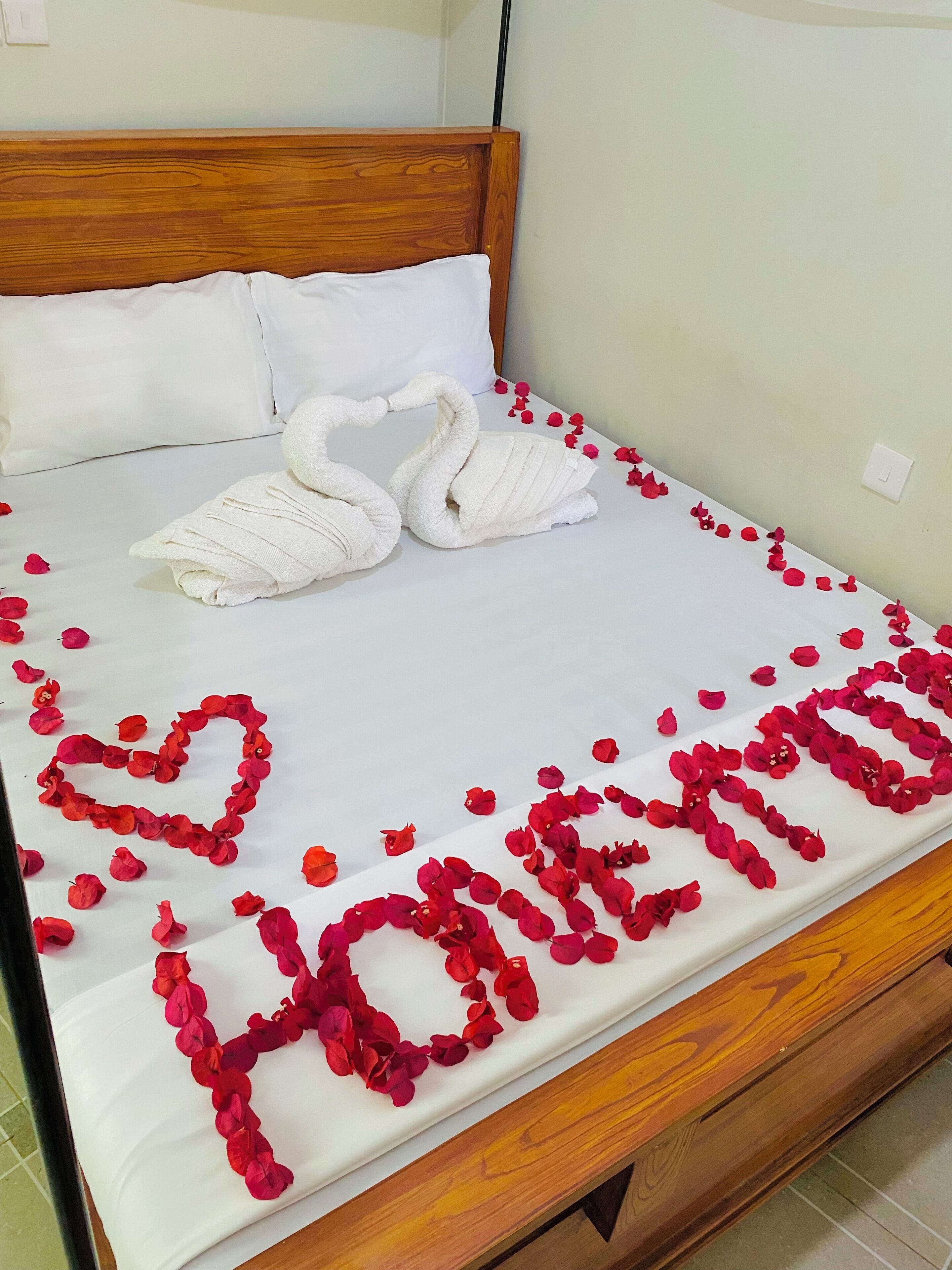 Una cama con sábana blanca, dos toallas blancas dobladas en forma de cisnes, y pétalos de flores rojas dispuestos para deletrear 'Love' sobre la cama.