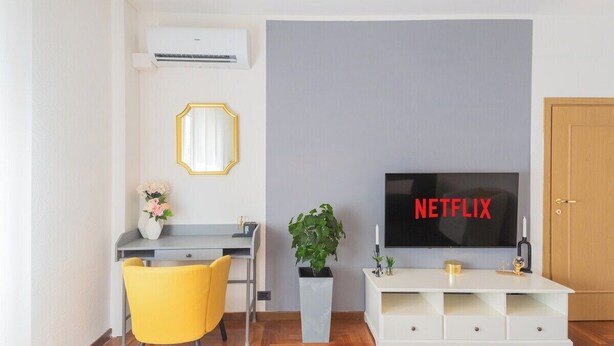 A modern living room with a grey wall, a white entertainment unit, a yellow chair, a desk, a potted plant, and a flat-screen TV displaying "NETFLIX".