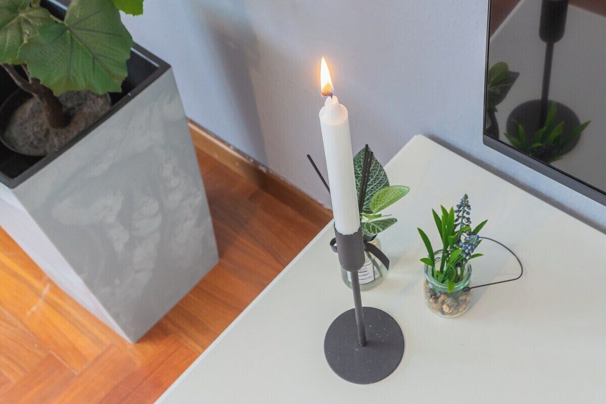 A white candle on a black holder on a white table, with a potted plant and a small glass container with a plant on the table.