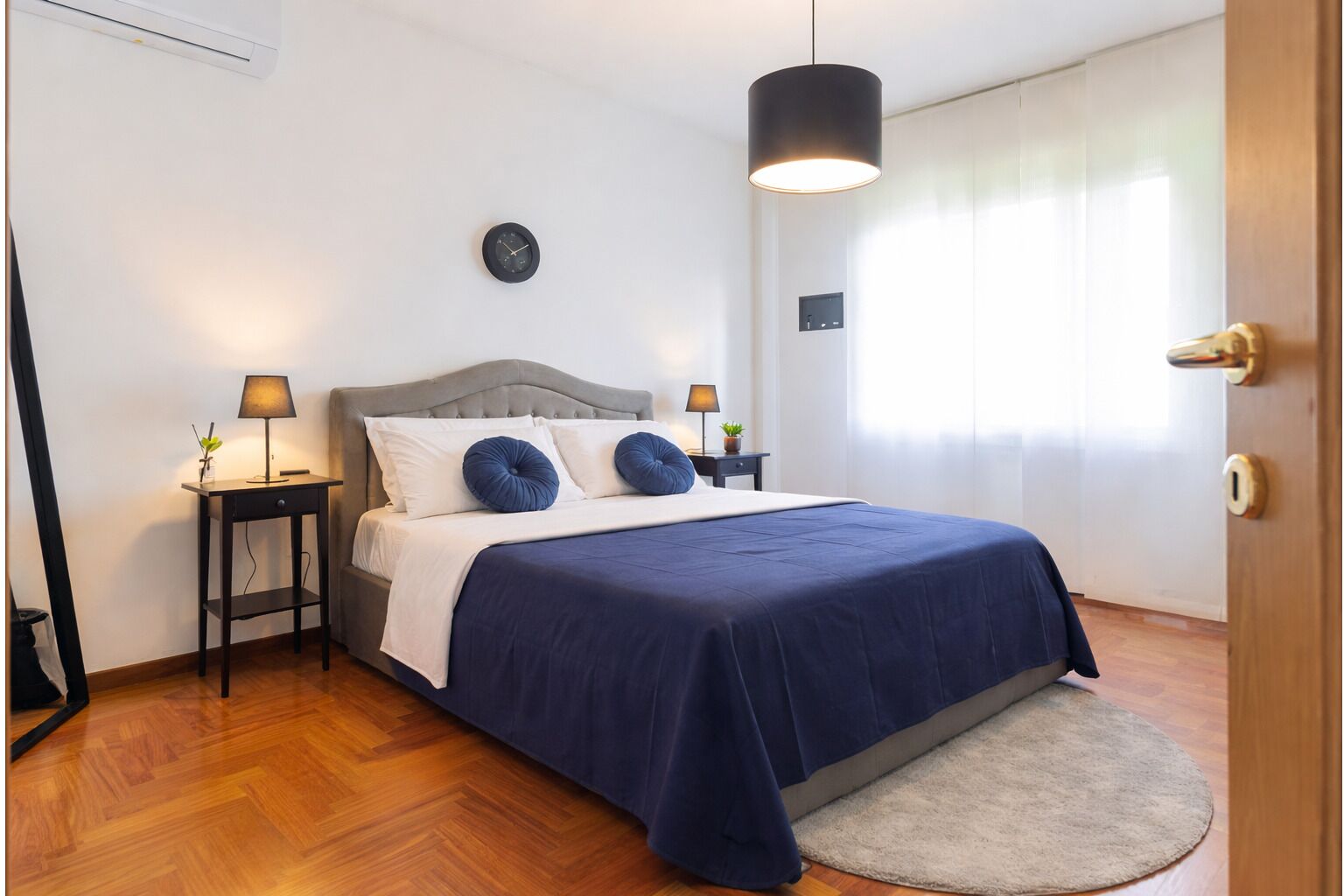 Une chambre à coucher bien rangée, avec un lit, deux tables de chevet, une horloge murale, un luminaire suspendu et une fenêtre avec des rideaux.