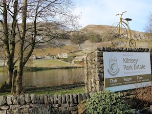 Cartel de Kilnsey Park Estate mostrando una vista panorámica de un lago, un muro de piedra y una bicicleta.