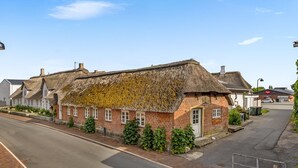 Ein traditionelles Haus mit Strohdach und Backsteinfassade, einem kleinen Vorbau und einer schmalen Straße im Hintergrund.