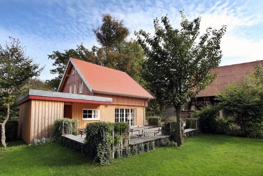 Une maison en bois avec un toit rouge, entourée d’un jardin bien entretenu et d’arbres.