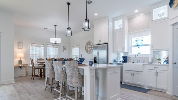 A modern kitchen with white cabinetry, a central island, pendant lighting, and a dining area with wicker chairs. - Indian Sunrise- peaceful/short stroll to beach! (Galveston)