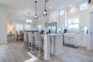 A modern kitchen with white cabinetry, a central island, pendant lighting, and a dining area with wicker chairs. - Indian Sunrise- peaceful/short stroll to beach! (Galveston)