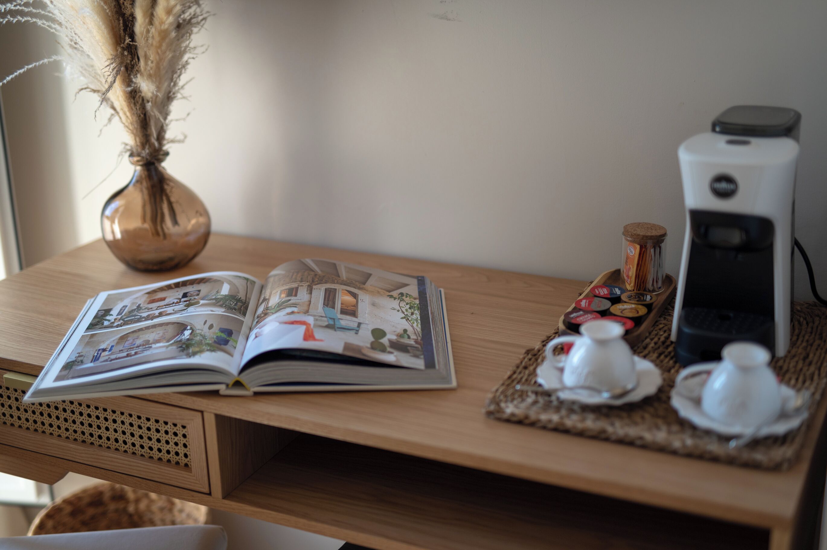 A wooden table with a magazine, a vase with dried plants, a coffee maker, and a tray with teacups and saucers.