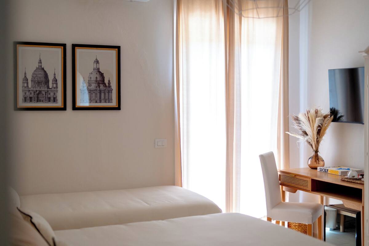 Hotellihuone, jossa on kaksi sänkyä, työpöytä tuolilla, maljakko, jossa on koristeheinää, ja kaksi kehystettyä kuvaa seinällä.