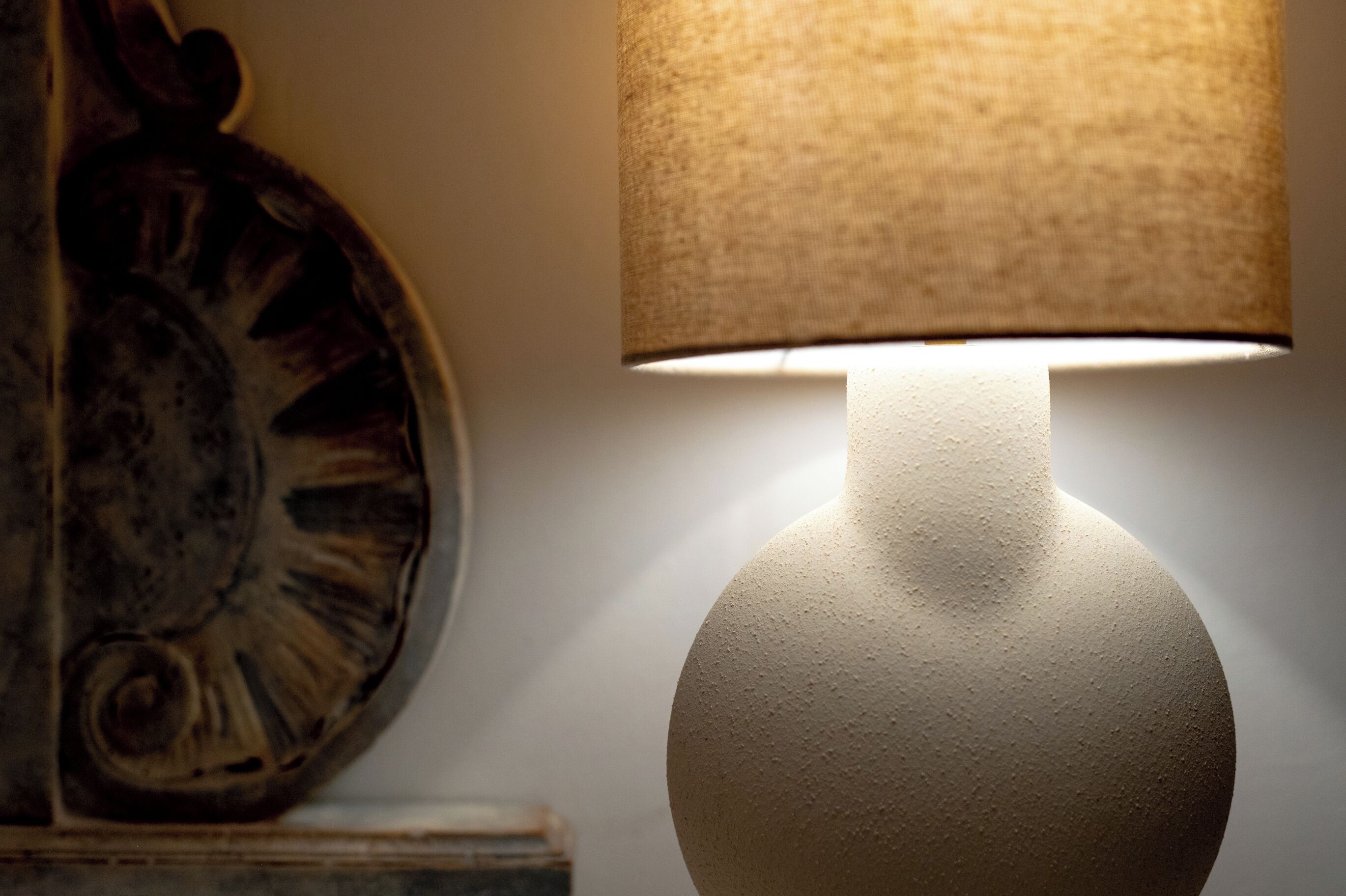 Una lampada con un paralume lavorato e una base rotonda, posizionata accanto a un muro con un piatto decorativo.