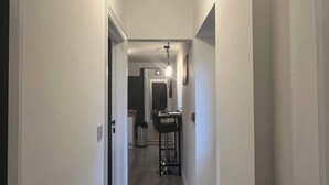 Um corredor estreito com uma lâmpada no teto, uma porta à esquerda e uma área de cozinha ao longe.