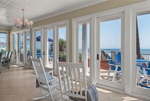 Eine überdachte Terrasse mit weißen Schaukelstühlen und blauen Sesseln, große Fenster mit Blick auf den Ozean und eine Kristallleuchter.