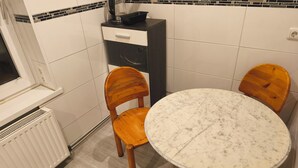 Eine kleine Küche mit weißen Fliesen, einem weißen Heizkörper, einem schwarz-weißen Schrank und einem runden Marmortisch mit vier Holzkästen.