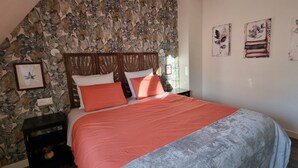 A bedroom with a sloped ceiling, a bed with a red and grey bedspread, a wooden headboard, and a stone-patterned wall. There are framed pictures on the wall and a small nightstand with a lamp. - Au Pré du Chêne (Le Faouët)