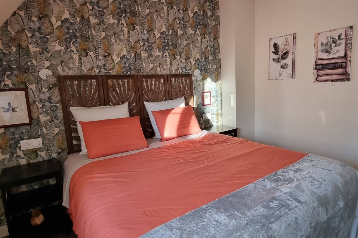 A bedroom with a sloped ceiling, a bed with a red and grey bedspread, a wooden headboard, and a stone-patterned wall. There are framed pictures on the wall and a small nightstand with a lamp.