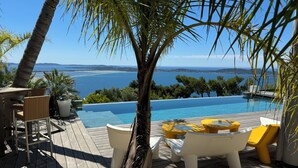 A poolside area with a view of the sea, wooden deck, white lounge chairs, and yellow tables. - 180°C sea Giens peninsula (Carqueiranne)