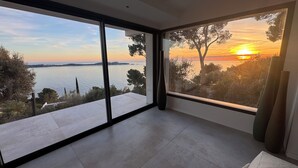 A room with large floor-to-ceiling windows offering a view of a sunset over a body of water, surrounded by trees. - 180°C sea Giens peninsula (Carqueiranne)