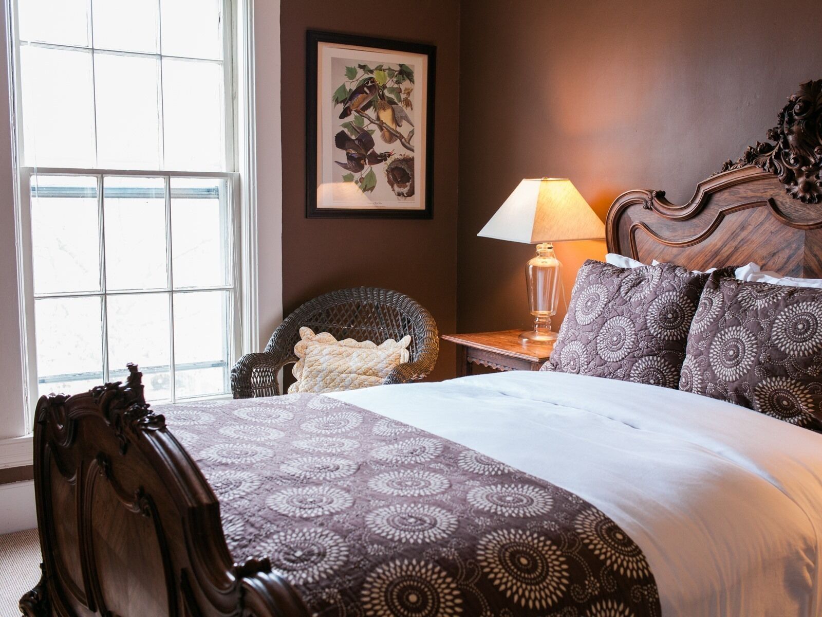 A bedroom with a large window, a wooden headboard, a patterned bedspread, a nightstand with a lamp, and a wicker chair.