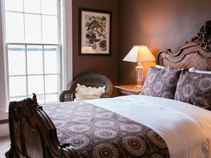 Une chambre avec une grande fenêtre, une tête de lit en bois, une couvre-lit à motifs, une table de chevet avec une lampe et un fauteuil en osier.
