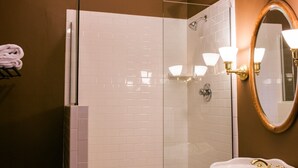 Um banheiro com chuveiro em azulejos brancos, pia com detalhes dourados e espelho com moldura de madeira.