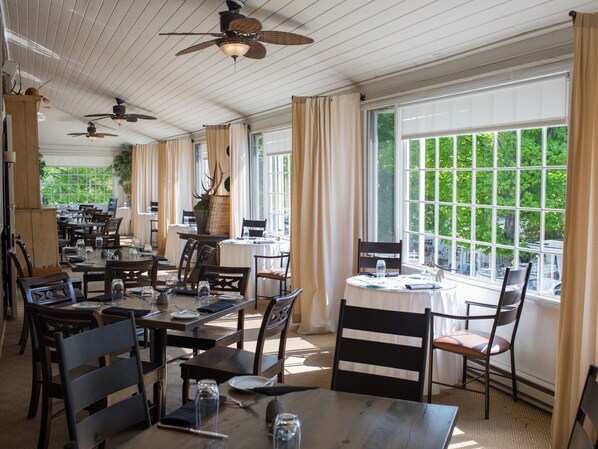 Un restaurant doté de nombreuses tables et chaises, de ventilateurs de plafond, de grandes fenêtres avec des rideaux et offrant une vue sur la verdure environnante.