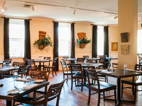 A restaurant with multiple tables and chairs, large windows with curtains, and wall-mounted baskets with plants.