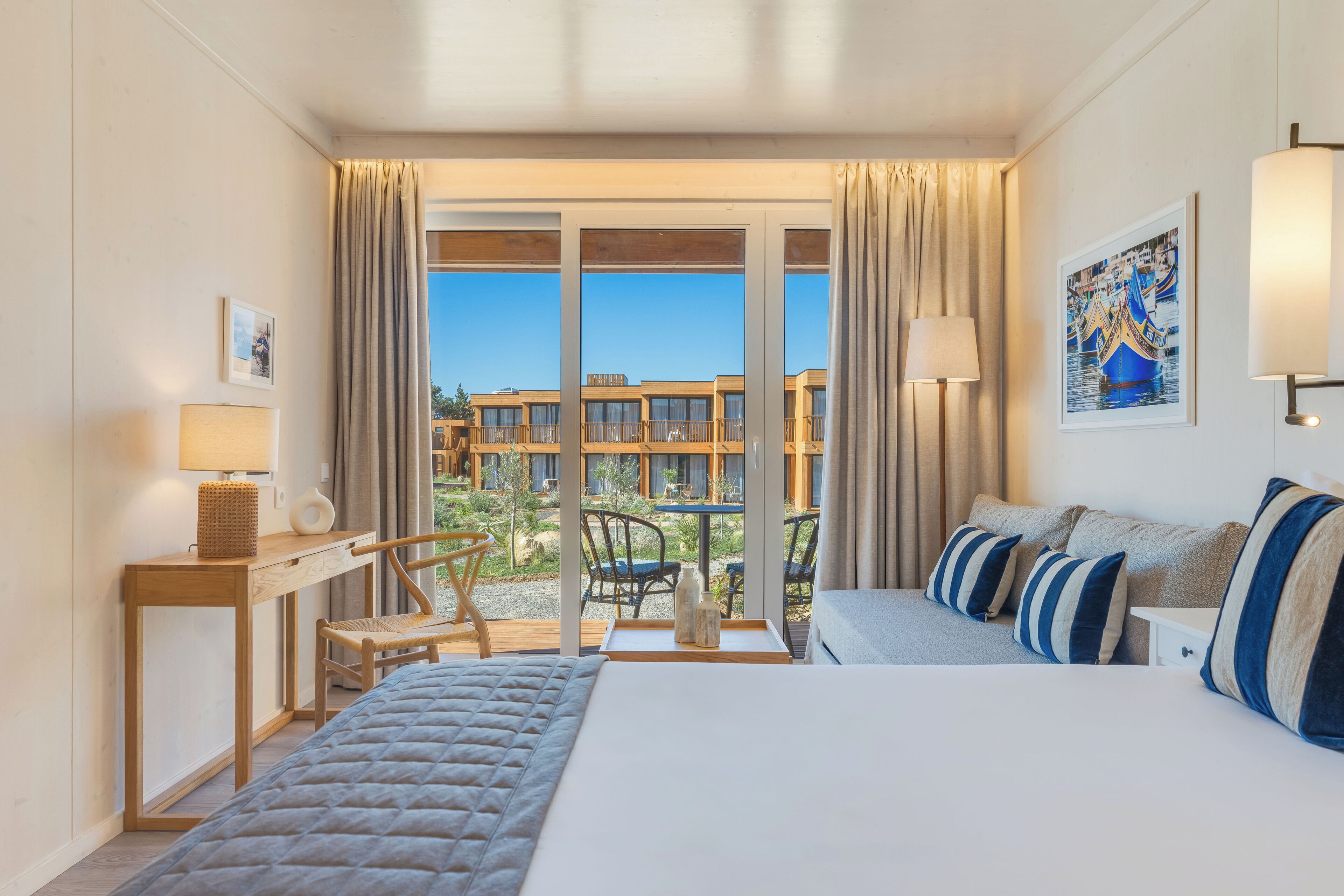 A hotel room with a bed, a desk, a chair, a balcony with a view of buildings, and a painting on the wall.