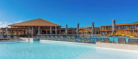 A clear swimming pool with lounge chairs and umbrellas, a wooden pavilion, and a building with balconies.