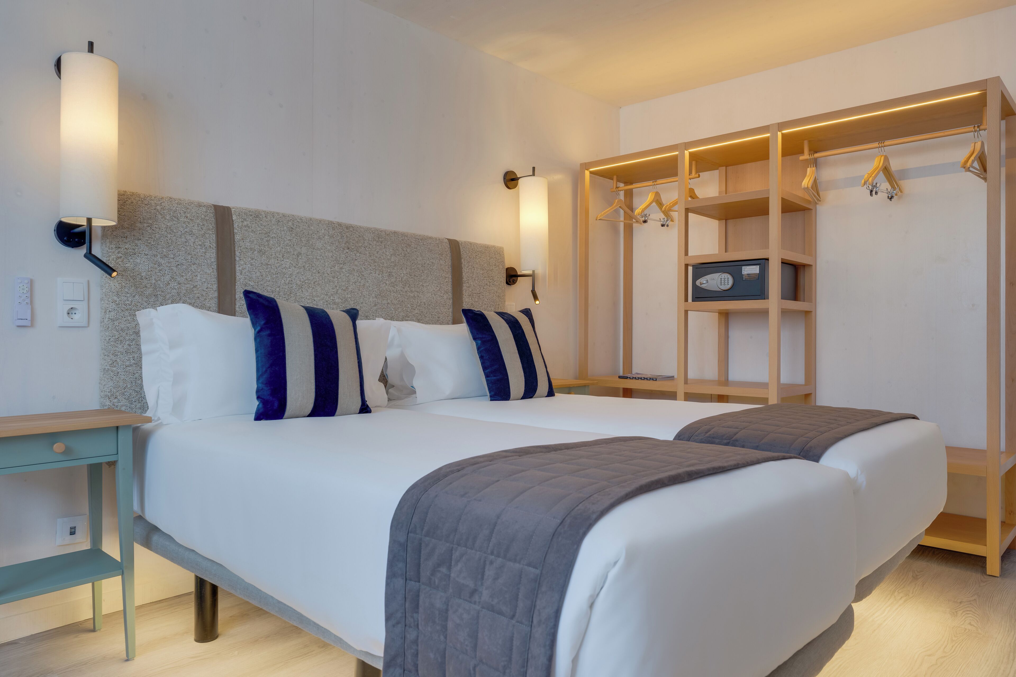 Una cama doble con colcha gris, una mesita de noche de madera, una lámpara de pared y un armario de madera con espacio para colgar.