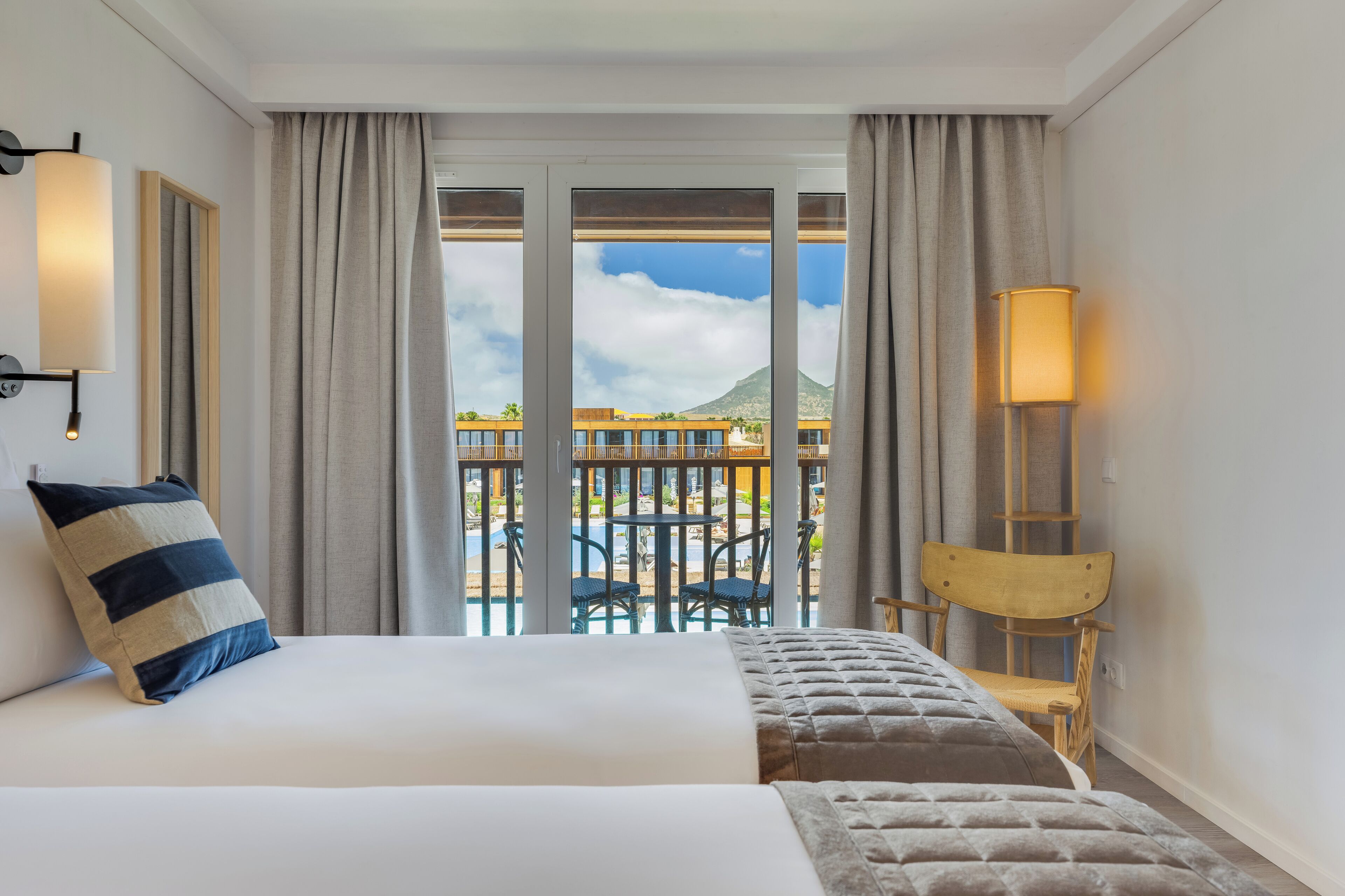A hotel room with a bed, a chair, a table, a lamp, and a balcony with a view of a mountain and pool.