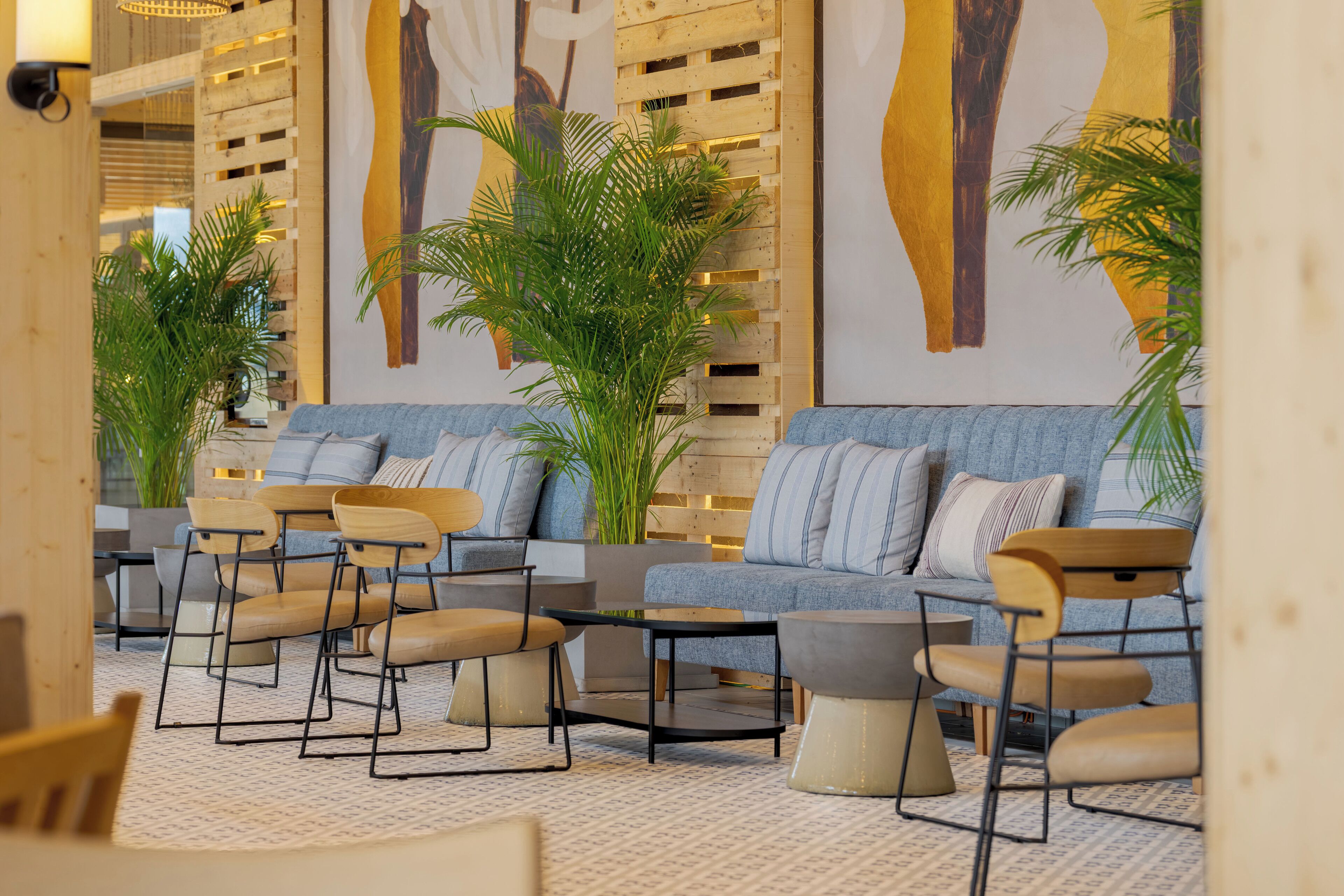 Un área de descanso moderna con sofás, sillones y una mesa de centro, con una pared de madera decorada con un mural de un árbol y plantas en macetas.
