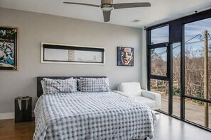 Ein modernes Schlafzimmer mit einem großen Bett, einem weißen Sessel und Blick ins Freie durch große Fenster.