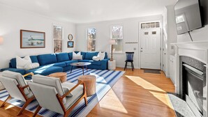 Een woonkamer met een blauwe modulaire bank, een ronde salontafel en twee fauteuils. Er is een open haard, een aan de muur bevestigde televisie en een deur naar een andere kamer.