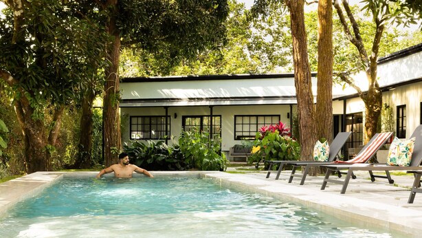 Una piscina con una persona nadando, rodeada de sillas de descanso y un edificio con ventanas y plantas.