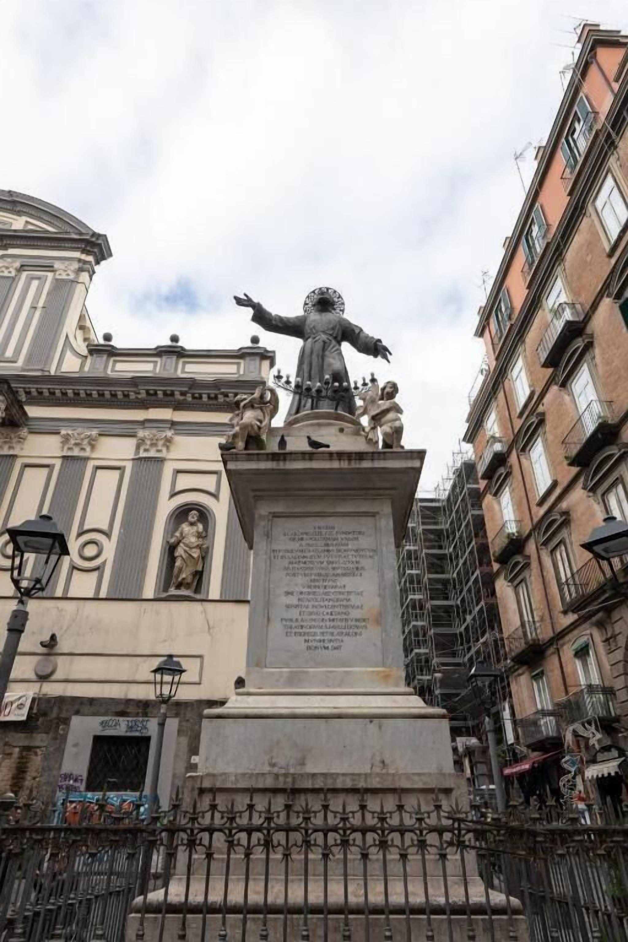 Una statua con iscrizioni su un piedistallo, circondata da un recinto in ferro battuto, con edifici sullo sfondo.