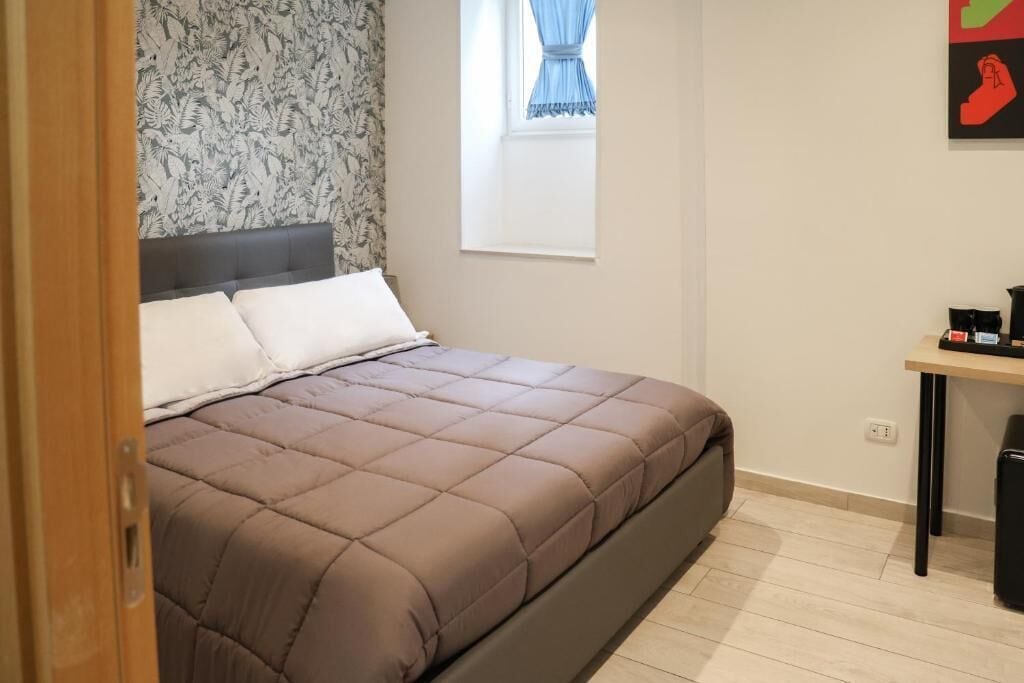 A bed with a headboard, a window with curtains, a small table with a coffee maker, and a patterned wallpaper.